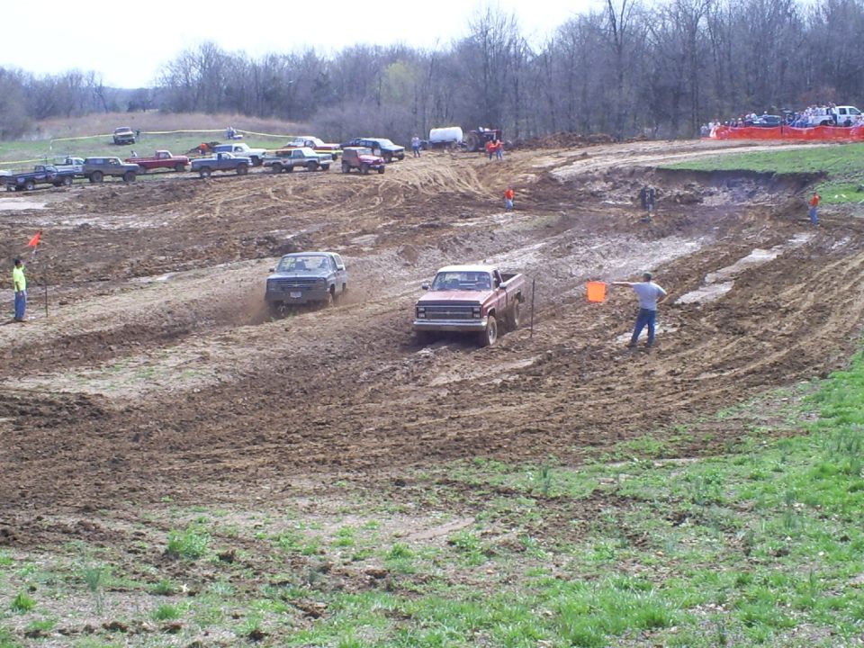 Two pickup trucks racing through mud bog lanes at an off-road event with flagman and spectators