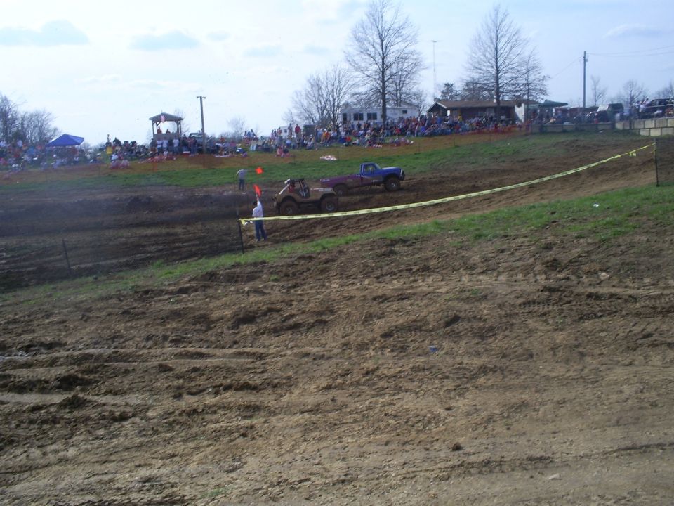 Blue pickup and Jeep at the line of a dirt mud race with crowd behind