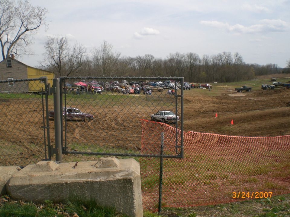 Maroon pickup and silver sedan on a dirt mud-race track behind chain-link and orange fence