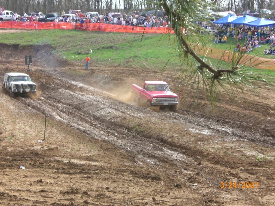 Red pickup slinging mud down a rutted mud-drag pit with crowd and canopies behind