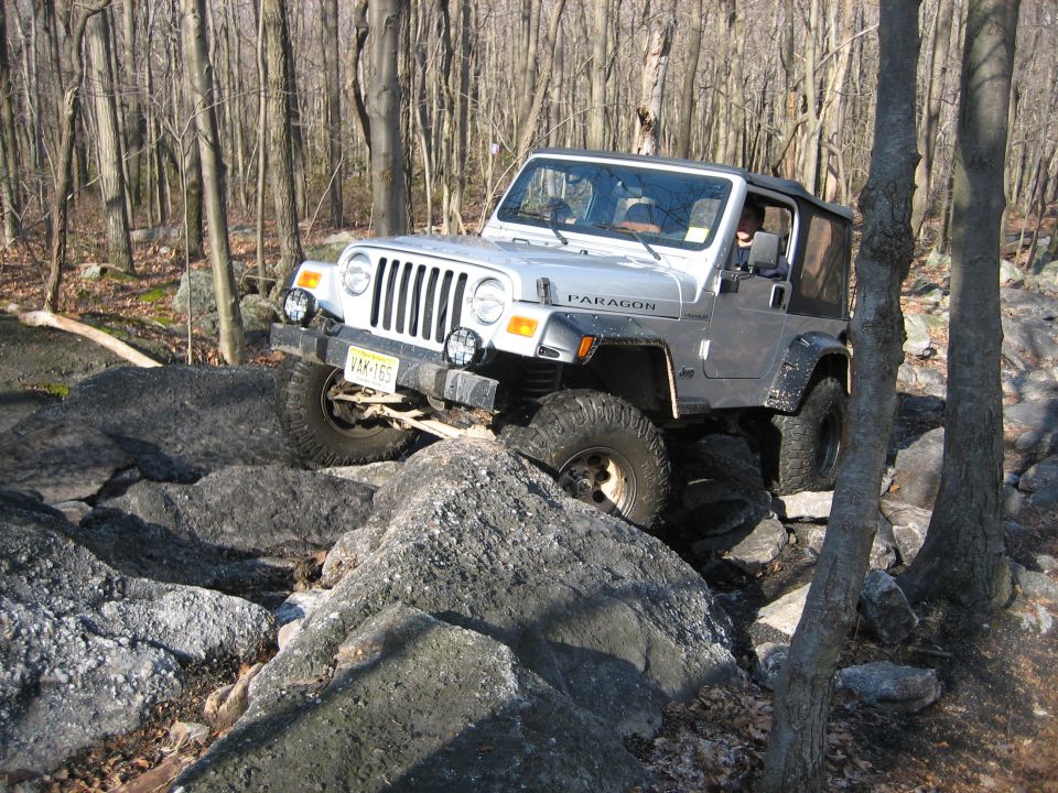 Silver Jeep Wrangler navigating a rocky trail in a wooded area. License plate is visible