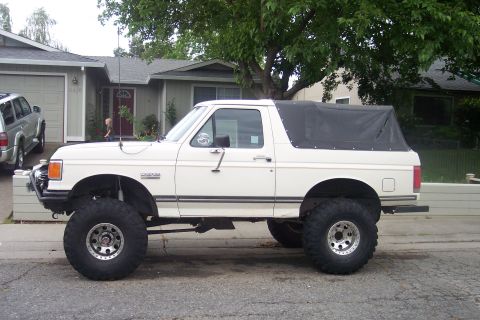 1989 Ford Bronco - This lifted Ford Bronco has a removable soft top and is parked on a residential street next to a silver SUV.