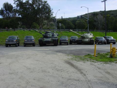 1994 Ford Bronco - A lineup of military tanks and SUVs are parked on a gravel lot with trees and traffic lights in the background.