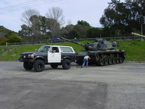 1994 Ford Bronco - A Ford Bronco tows a military tank down a paved road with a person attending to the connection.