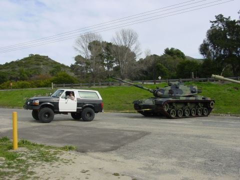 1994 Ford Bronco - A two-tone Ford Bronco is seen alongside a military tank on a paved surface with grassy hills in the background.