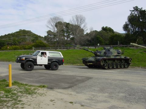 1994 Ford Bronco - A two-tone Ford Bronco is parked on asphalt next to a military tank.