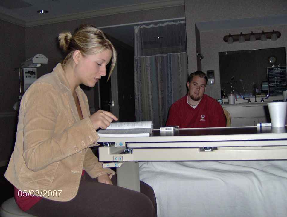 Woman in tan jacket points at book, man in red shirt sits behind table with cup