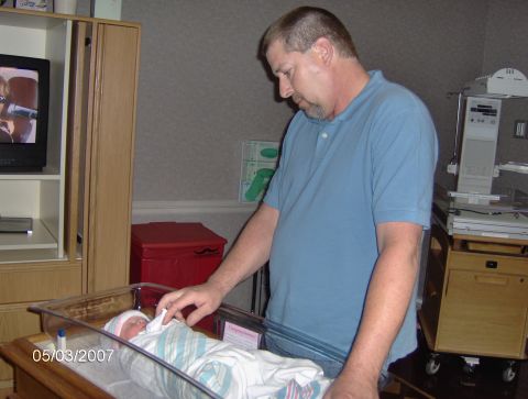 1978 Ford Bronco - A man gently touches a newborn baby in a bassinet, with medical equipment in the background.