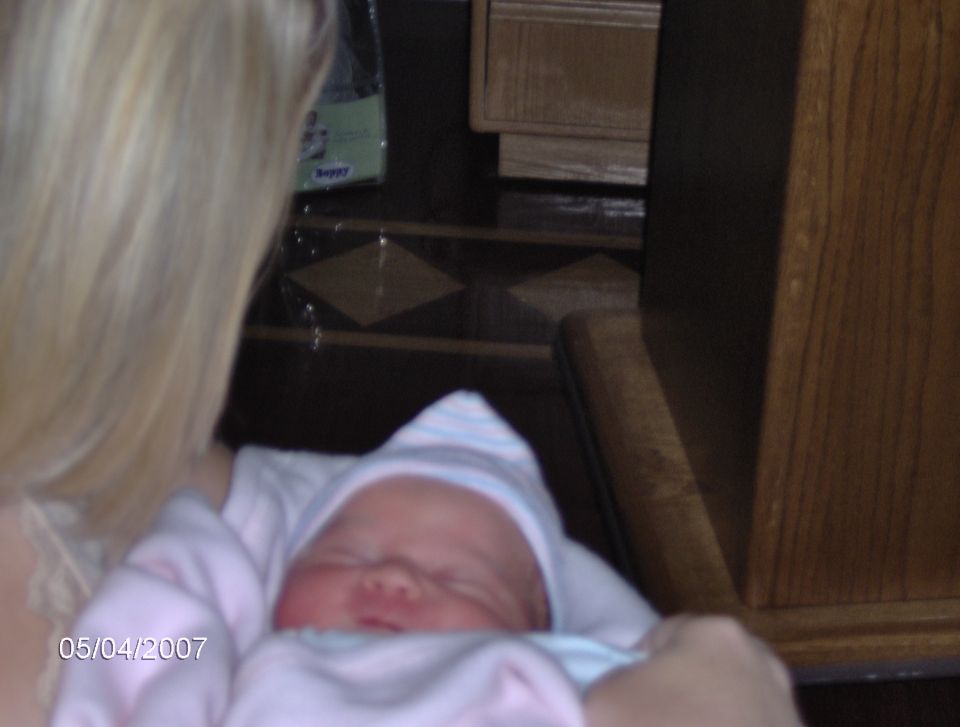 Newborn baby in a pink and blue striped hat and blanket, held by a woman with blonde hair. Date stamp reads 05/04/2007