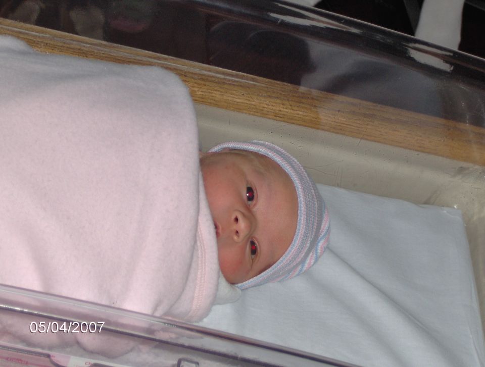 Newborn baby in a hospital bassinet, wrapped in a pink blanket, wearing a striped hat. Date stamp reads 05/04/2007