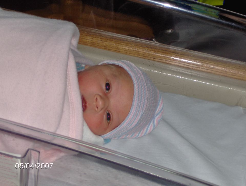 Newborn baby wrapped in pink blanket, wearing a striped hat, in a hospital bassinet. Date stamp reads 05/04/2007