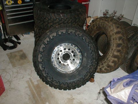 1996 Ford Bronco - A mud-terrain tire mounted on a polished aluminum beadlock wheel with a bolted outer ring, standing on a garage floor in front of a black t…