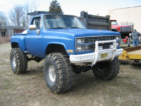 1990 Ford Bronco - A lifted blue pickup truck with large off-road tires sits on a grassy lot next to a trailer and another truck.