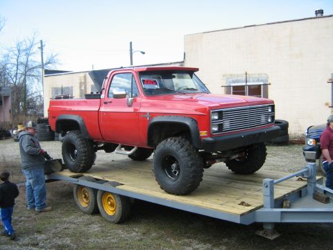 1990 Ford Bronco - Red pickup truck with large tires loaded onto a trailer. A "FOR SALE" sign is visible in the driver's side window.