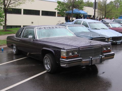 1998 GMC Sierra 1500 - A dark Cadillac is parked in a lot on a rainy day, with text on the windshield and other vehicles nearby.