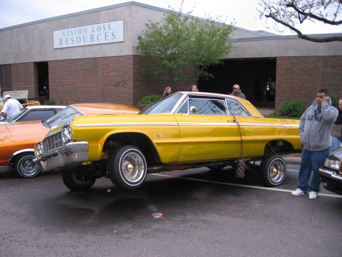 1998 GMC Sierra 1500 - A yellow 1964 Chevrolet Impala lowrider is parked next to an orange car in front of a building.