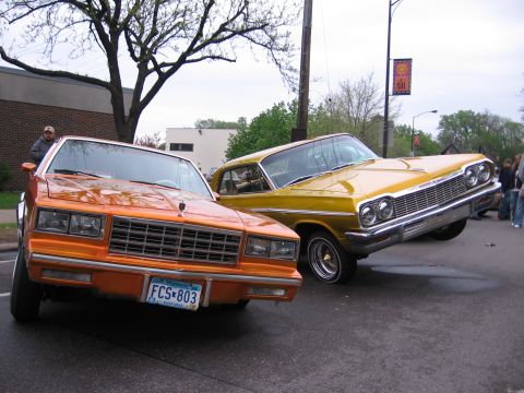 1998 GMC Sierra 1500 - Two custom cars, an orange and a yellow lowrider, are parked on a street during an outdoor event.