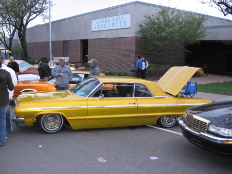 1998 GMC Sierra 1500 - A yellow 1964 Chevrolet Impala lowrider is parked with its trunk open, alongside other vehicles.
