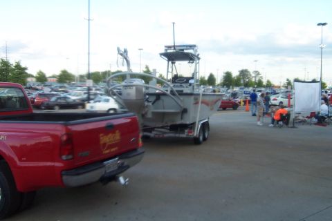 1998 Ford Crown Victoria - A red pickup truck tows a boat on a trailer through a crowded parking lot with other vehicles and people visible.