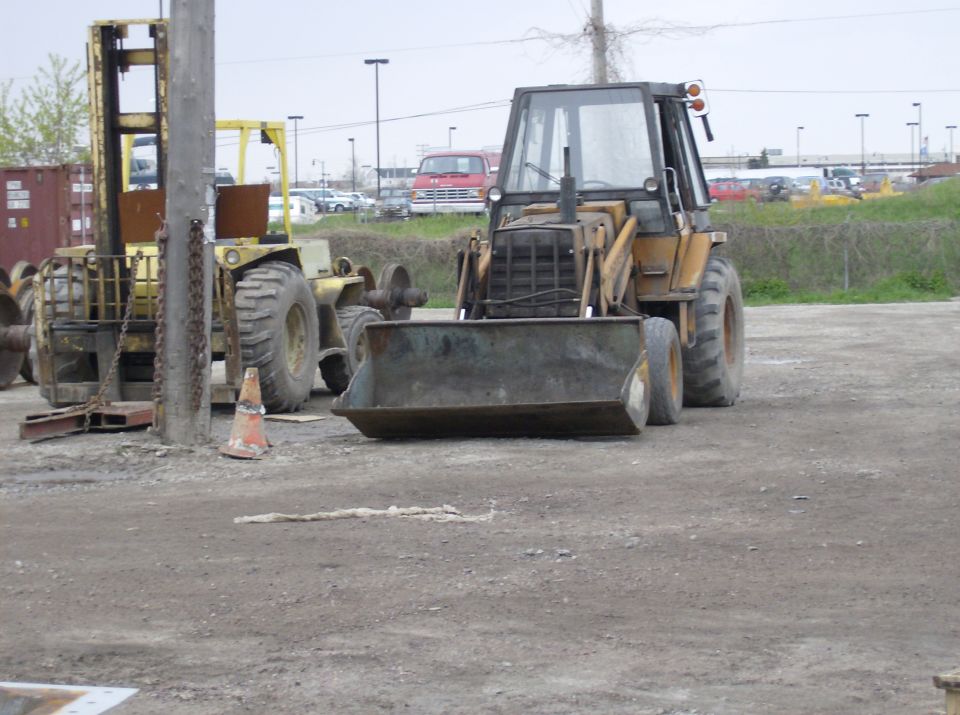 Yellow backhoe loader with front bucket and cab, next to a yellow forklift. Construction site setting