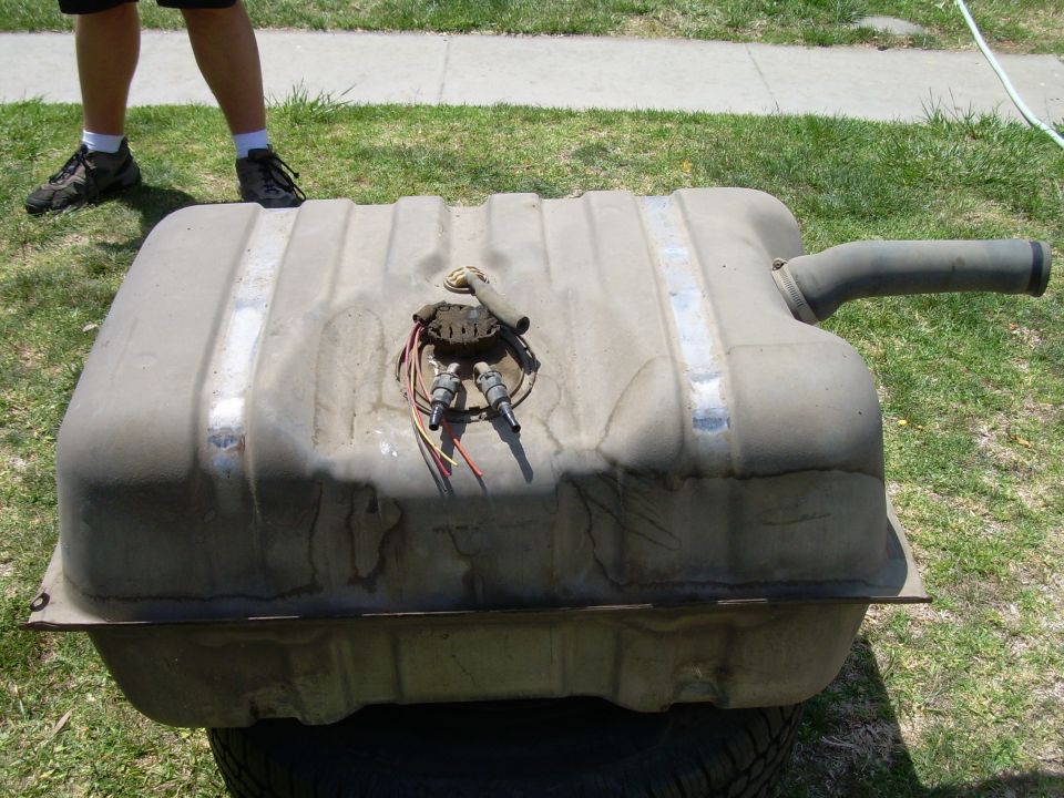 Fuel tank with fuel pump assembly and filler neck removed, resting on a tire outdoors