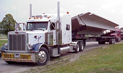1979 Ford Bronco - A white Peterbilt semi-truck transports a massive concrete beam, with another red truck visible behind it.