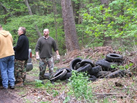 1996 Ford Bronco - A silver SUV is parked near a pile of discarded tires in a wooded area, with several people standing nearby.