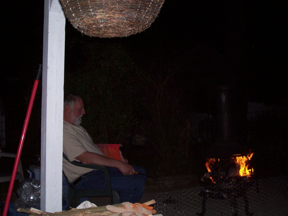 Man sits in a chair next to a fire pit at night, with a hanging basket overhead