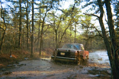 1990 Ford Bronco - A red pickup truck navigates a muddy trail through a forest.