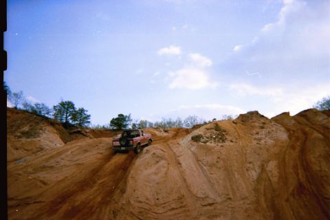 1990 Ford Bronco - Red 1990 Ford Bronco tackles a steep sandy incline under a partly cloudy sky.