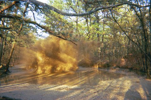 1990 Ford Bronco - A vehicle navigates a water-logged dirt path, creating a dramatic spray of mud and dust amidst the trees.