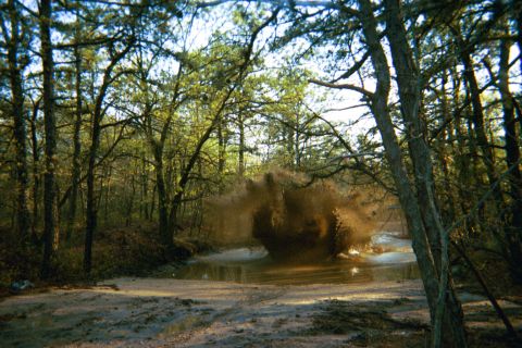 1990 Ford Bronco - An off-road vehicle creates a large splash as it drives through a muddy puddle in a forest.