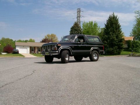 1984 Ford Bronco - A black Ford Bronco is parked on a street in front of houses and trees under a blue sky.