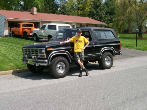 1984 Ford Bronco - A man poses with a black Ford Bronco, while two other trucks are visible in the background.