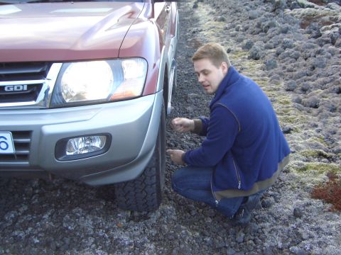 1974 Ford Bronco - Man checks tire on a Mitsubishi Pajero SUV. The vehicle is parked on a rocky, mossy terrain.