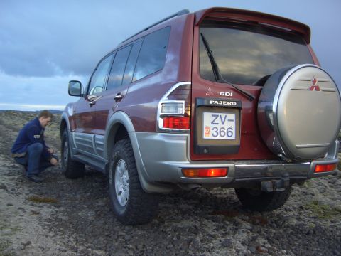 1974 Ford Bronco - A person kneels by the rear tire of a red Mitsubishi Pajero SUV parked on a rocky, desolate landscape.