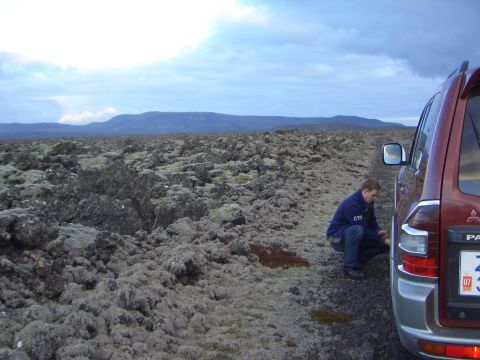 1974 Ford Bronco - A man checks the tire of a red SUV parked on a desolate, rocky landscape under a cloudy sky.