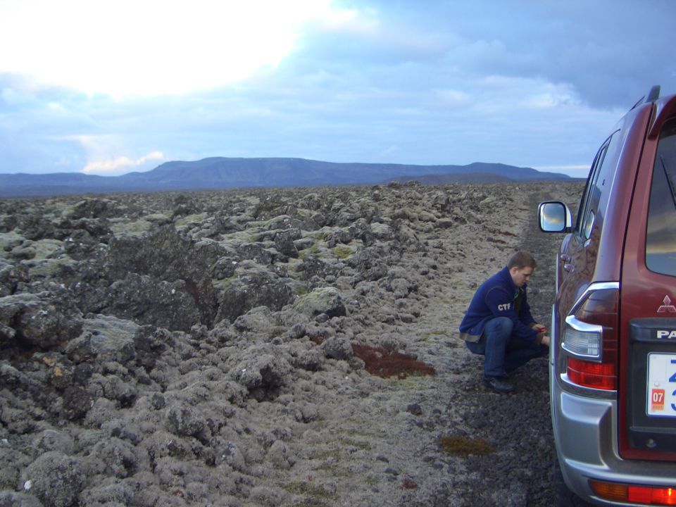 Red SUV parked on a rocky, moss-covered terrain with a man checking the tire. License plate visible