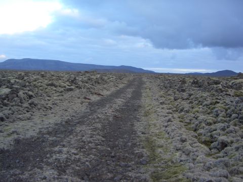 1974 Ford Bronco - A remote dirt track winds through a moss-covered, rocky terrain with distant mountains under a dramatic sky.