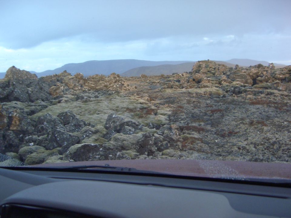 Rocky, moss-covered terrain under a cloudy sky, viewed from inside a vehicle