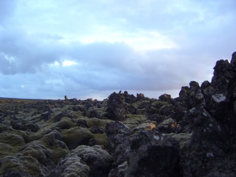1974 Ford Bronco - A vast lava field stretches under a dramatic sky, with jagged rocks covered in moss dominating the foreground.
