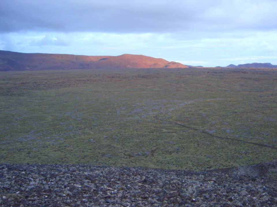 Wide view of a moss-covered plain with distant, sunlit mountains under a cloudy sky