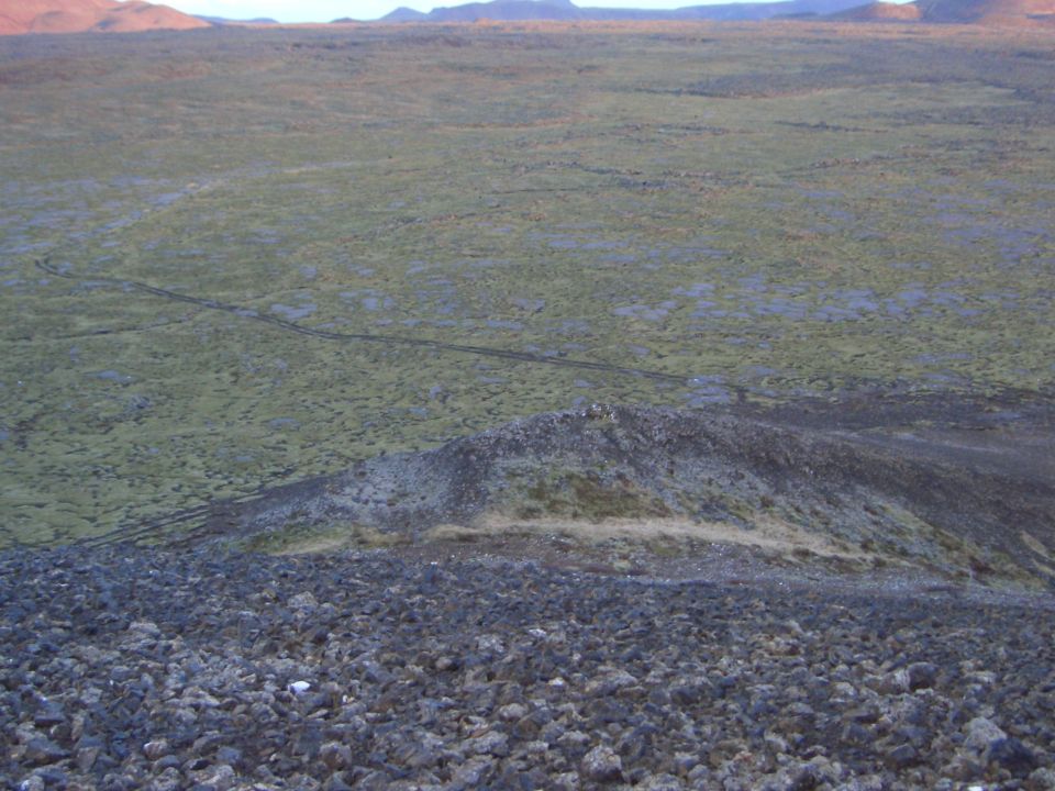 Rocky foreground slopes down to a vast, green, moss-covered plain with tire tracks, under a hazy sky
