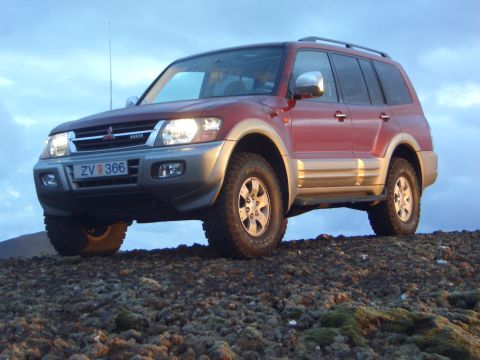 1974 Ford Bronco - A red Mitsubishi Pajero SUV is parked on a rocky surface with a cloudy sky in the background.