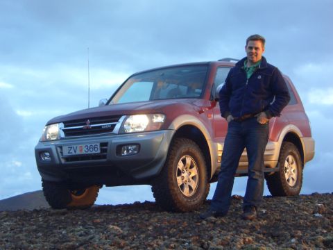 1974 Ford Bronco - A man poses next to a red Mitsubishi Pajero SUV on a rocky landscape under a cloudy sky.
