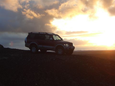 1974 Ford Bronco - A red SUV is positioned on a dark, rocky terrain as the sun sets in the background.