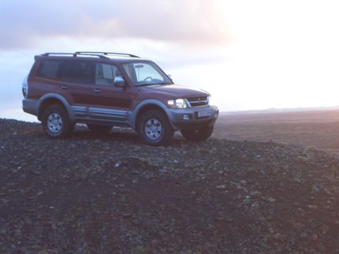 1974 Ford Bronco - A red SUV with a roof rack is parked on a rocky surface under a cloudy sky.
