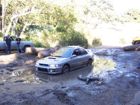 1991 Ford Bronco - A Subaru Impreza is stuck in a large mud puddle during an off-road event.