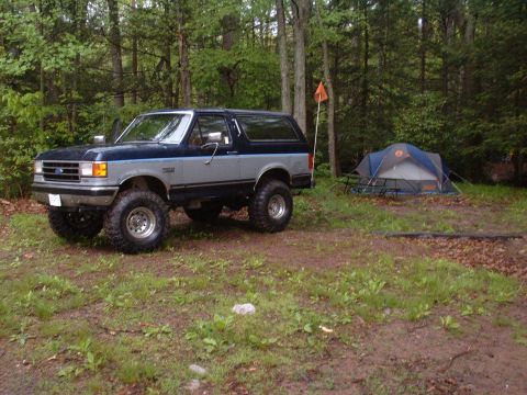 1991 Ford Bronco - A lifted Ford Bronco sits parked in a wooded area beside a campsite.