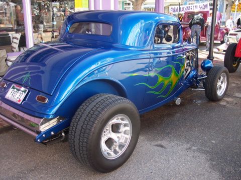 1972 Ford Ranchero - A custom blue hot rod with flame graphics is displayed at a car show, with other vehicles visible in the background.
